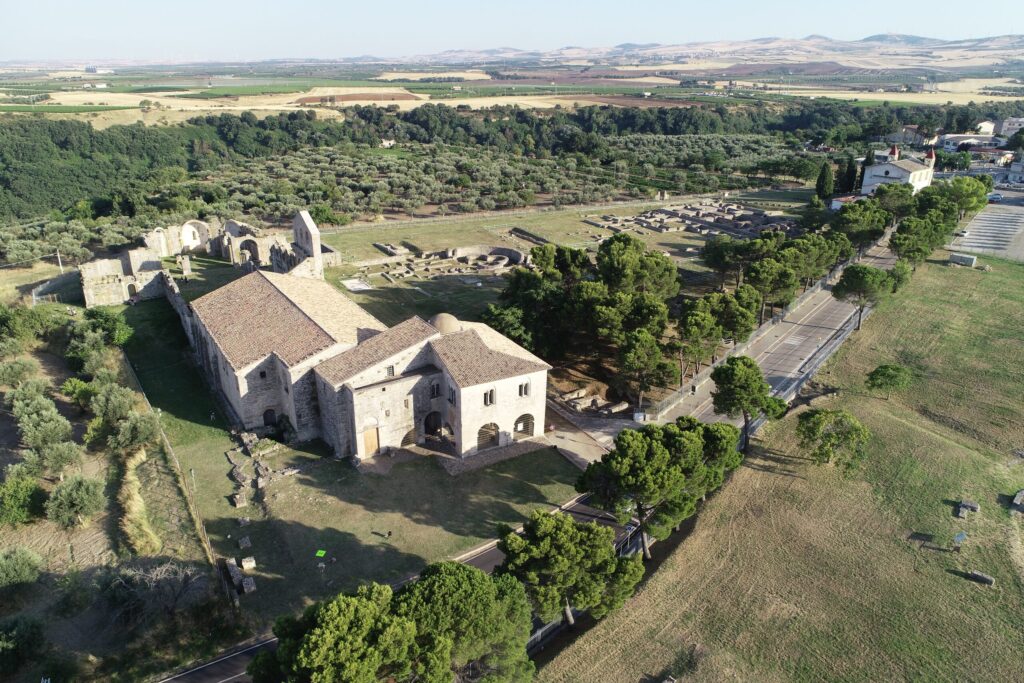 Vista aerea dell'abbazia della Santissima Trinità e delle rovine dell'Incompiuta tra campi di ulivi e aree archeologiche.