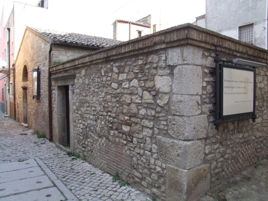 Edificio in pietra e mattoni con tetti in tegole, un vicolo acciottolato e un pannello esplicativo di Achille Cappellano.